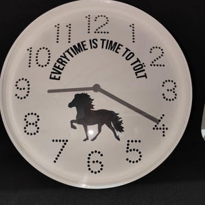 Könnte beinhalten: Weiße runde Uhr mit schwarzen Zahlen und Zeigern. Das Zifferblatt zeigt eine schwarze Pferdesilhouette und den Text "EVERYTIME IS TIME TO TÖLT". Die Uhr befindet sich vor einem schwarzen Hintergrund.