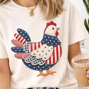May include: Cream-colored t-shirt featuring a patriotic chicken design. The chicken is adorned with red, white, and blue patterns, including stars and stripes, and has a red comb and beak. The design is a stitched applique.