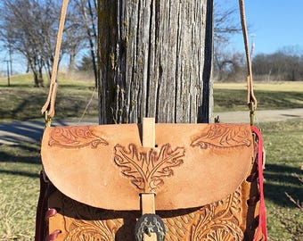 May include: A tan leather crossbody bag with intricate floral and leaf carvings. The bag features a flap closure with a buckle and red fringe along the bottom. The strap is a lighter tan color.