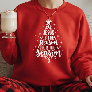 Könnte beinhalten: Ein rotes Sweatshirt mit dem weißen Text "Jesus is the Reason for the Season", gestaltet in Form eines Weihnachtsbaums. Die Person trägt eine rot-schwarz karierte Pyjamahose und hält ein Glas eines cremigen Getränks.