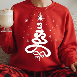 Könnte beinhalten: Ein rotes Sweatshirt mit einem weißen Weihnachtsbaum-Design, das "Jesus" schreibt. Das Design beinhaltet einen Stern und Schneeflocken. Die Person trägt eine rot-schwarz karierte Hose.