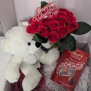 May include: A white teddy bear, a bouquet of red roses with a "Happy Valentine's Day" sign, and a box of Lindor milk chocolate truffles in a white gift box with red and white shredded paper.