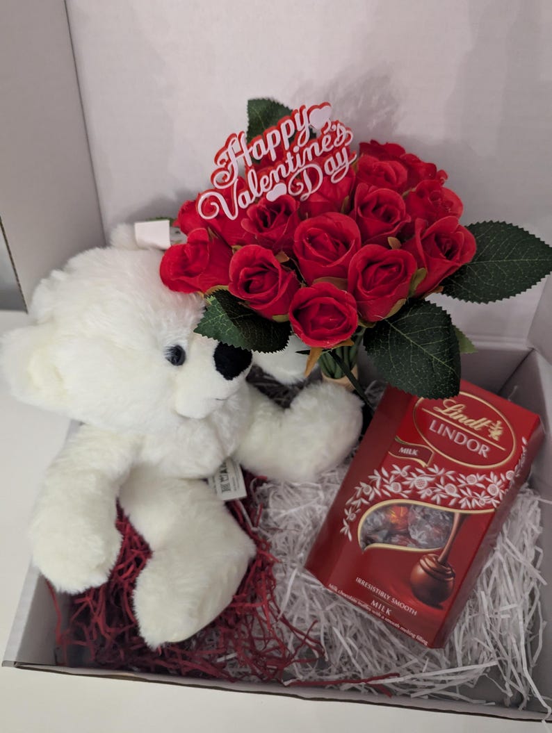 May include: A white teddy bear, a bouquet of red roses with a "Happy Valentine's Day" sign, and a box of Lindor milk chocolate truffles in a white gift box with red and white shredded paper.