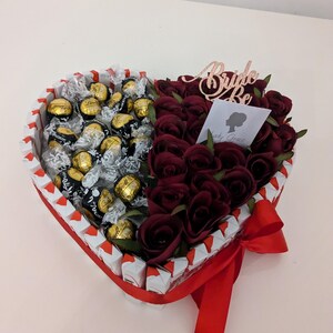 May include: A heart-shaped box filled with chocolate truffles and red roses. The box is decorated with a red ribbon and a sign that says "Bride to Be".