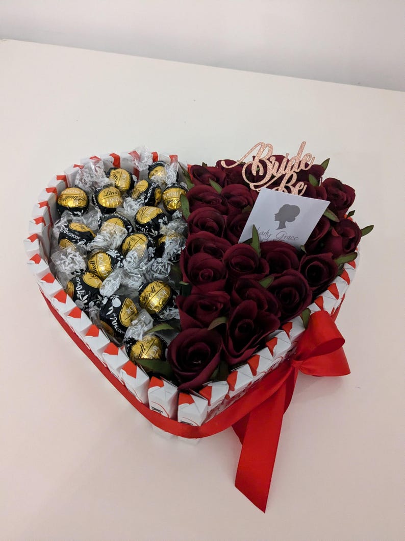 May include: A heart-shaped box filled with chocolate truffles and red roses. The box is decorated with a red ribbon and a sign that says "Bride to Be".