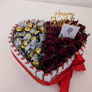 May include: A heart-shaped arrangement filled with chocolate truffles and red roses. The arrangement is decorated with a red ribbon and a gold "Happy Birthday" topper.