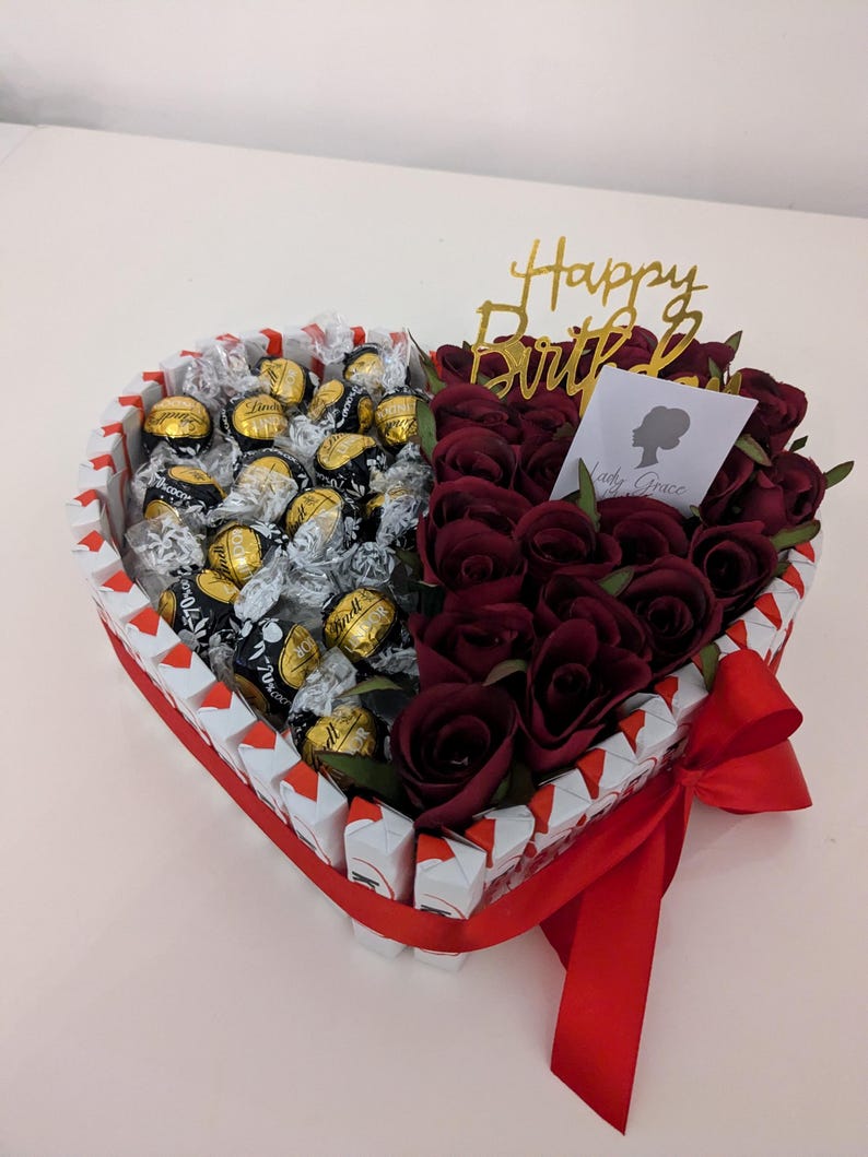 May include: A heart-shaped arrangement filled with chocolate truffles and red roses. The arrangement is decorated with a red ribbon and a gold "Happy Birthday" topper.