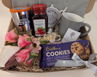 May include: A gift basket filled with a variety of items, including a jar of strawberry jam, a box of Cadbury milk chocolate cookies, a mug, a coffee packet, and a small bouquet of pink flowers.