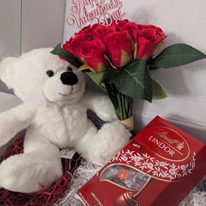 May include: A white teddy bear, a bouquet of red roses with a "Happy Valentine's Day" sign, and a box of Lindt Lindor milk chocolate truffles in a gift box.