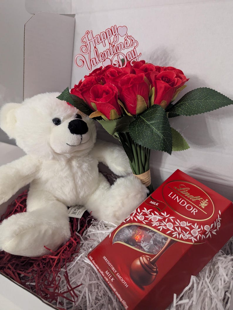 May include: A white teddy bear, a bouquet of red roses with a "Happy Valentine's Day" sign, and a box of Lindt Lindor milk chocolate truffles in a gift box.