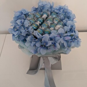 Puede incluir: Un ramo azul y blanco de hortensias artificiales con una caja de bombones Lindor en el centro.