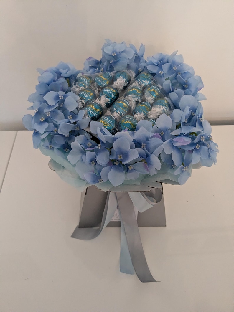 Puede incluir: Un ramo azul y blanco de hortensias artificiales con una caja de bombones Lindor en el centro.