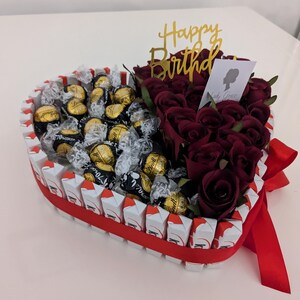 May include: A heart-shaped arrangement filled with chocolate truffles and red roses. The arrangement is decorated with a red ribbon and a gold "Happy Birthday" topper.