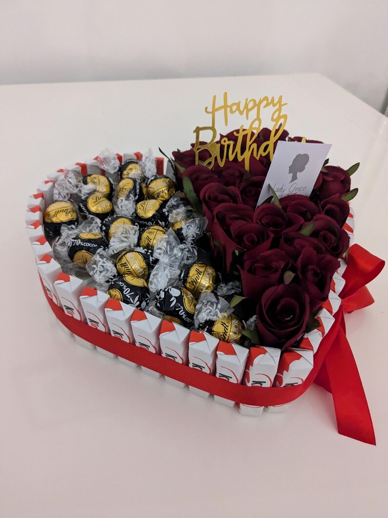 May include: A heart-shaped arrangement filled with chocolate truffles and red roses. The arrangement is decorated with a red ribbon and a gold "Happy Birthday" topper.