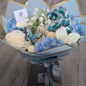May include: A blue and white floral arrangement in a box with a blue ribbon. The arrangement includes white roses, hydrangeas, and baby's breath. There are also chocolate truffles wrapped in blue foil.