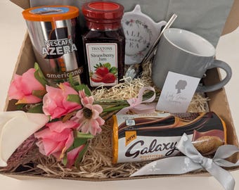 May include: A gift basket filled with a Nescafe Azera Americano coffee can, strawberry jam, a gray mug, a floral tea bag holder, a Galaxy Digestive Orange bar, and pink flowers. The items are arranged in a cardboard box with straw.