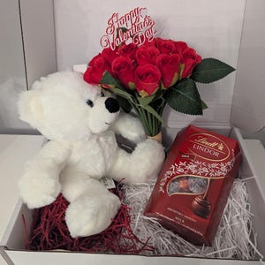 May include: A white teddy bear sits next to a bouquet of red roses with a "Happy Valentine's Day" sign. A box of Lindt Lindor milk chocolate truffles is also included in the gift box.