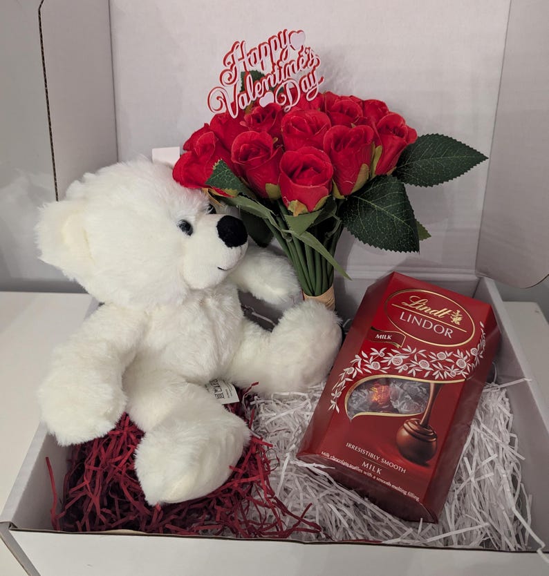 May include: A white teddy bear sits next to a bouquet of red roses with a "Happy Valentine's Day" sign. A box of Lindt Lindor milk chocolate truffles is also included in the gift box.