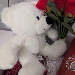 May include: A white teddy bear with black eyes and a black nose sits in a basket with red shredded paper. A bouquet of red roses is behind the bear.