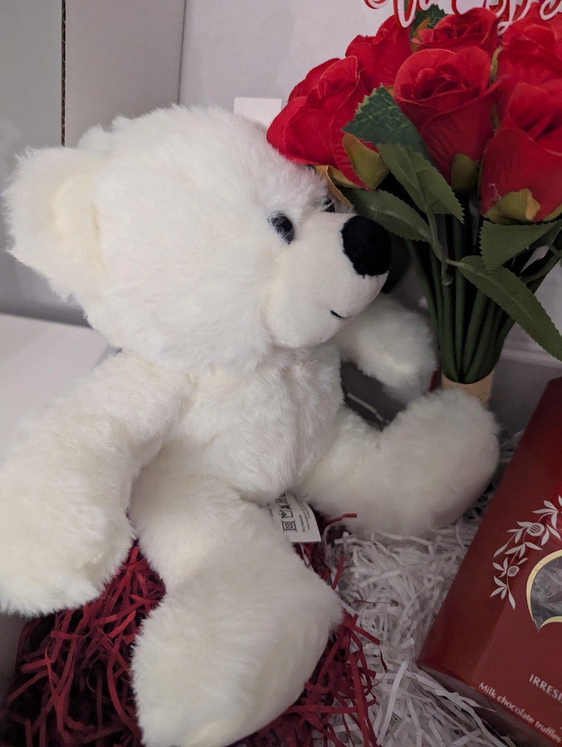 May include: A white teddy bear with black eyes and a black nose sits in a basket with red shredded paper. A bouquet of red roses is behind the bear.