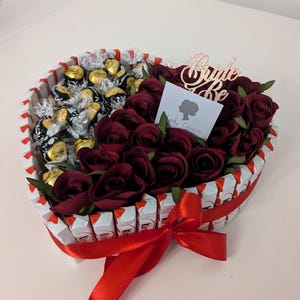 May include: A heart-shaped arrangement of chocolate candies and red roses, tied with a red ribbon. The arrangement is decorated with a gold glitter "Bride to Be" sign.