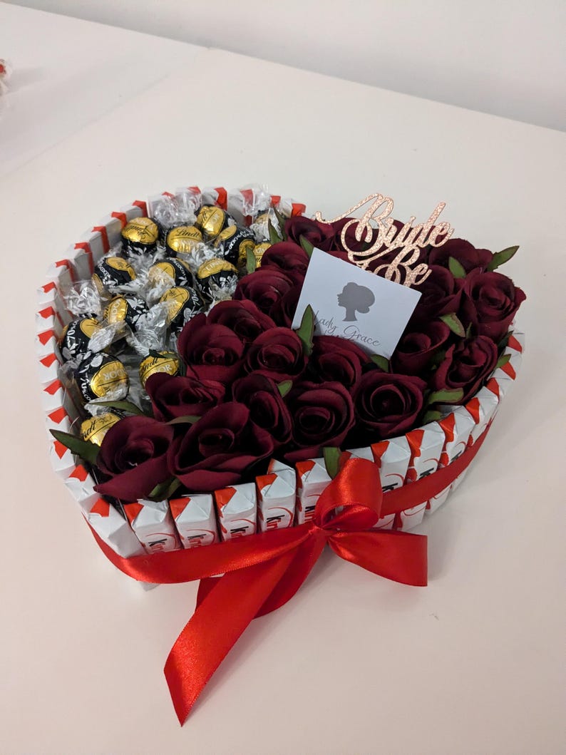 May include: A heart-shaped arrangement of chocolate candies and red roses, tied with a red ribbon. The arrangement is decorated with a gold glitter "Bride to Be" sign.