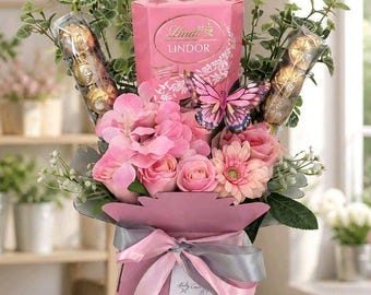 Puede incluir: Una caja de regalo rosa con un ramo de flores artificiales rosas, follaje verde, dos tubos transparentes de chocolates envueltos en oro y una caja de Lindt Lindor rosa. Incluye una mariposa decorativa y un lazo de color gris y rosa al frente.