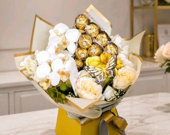 May include: A bouquet featuring white orchids, cream-colored roses, and gold-wrapped spherical chocolates, accented by a decorative butterfly. The arrangement is presented in a yellow box with a grey ribbon, labeled with Lady Grace Flowers text.