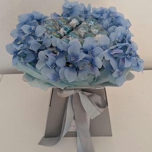 Puede incluir: Un ramo de hortensias azules arreglado en una caja gris con una cinta azul claro. El ramo est&aacute; lleno de chocolates envueltos en azul y verde.