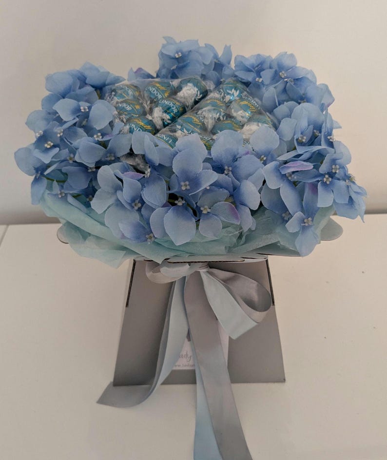 Puede incluir: Un ramo de hortensias azules arreglado en una caja gris con una cinta azul claro. El ramo est&aacute; lleno de chocolates envueltos en azul y verde.