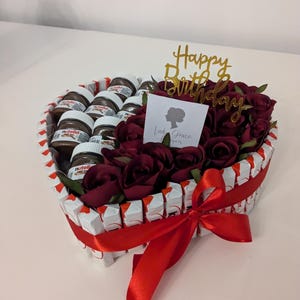 May include: A heart-shaped arrangement of chocolate spread jars and red roses with a red ribbon and a gold "Happy Birthday" topper.
