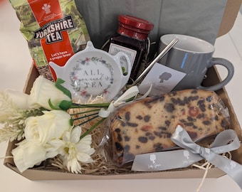May include: A gift basket filled with a white teacup with the text "All you need is tea", a mug, a jar of jam, a bag of Yorkshire tea, a loaf of fruitcake, white flowers, and a gray ribbon.
