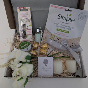 May include: A gift box filled with beauty and relaxation items. Includes a sheet mask, perfume, chocolates, candles, false eyelashes, and a lip balm. The box is decorated with a ribbon and a bouquet of white flowers. The box contains the text "Lady Grace Hampers".