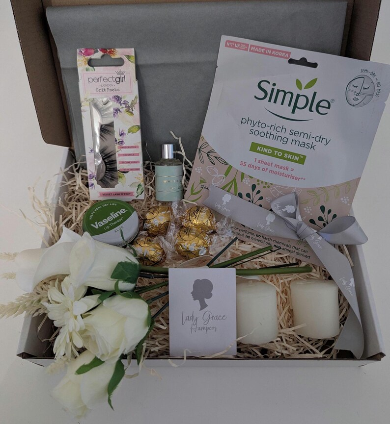 May include: A gift box filled with beauty and relaxation items. Includes a sheet mask, perfume, chocolates, candles, false eyelashes, and a lip balm. The box is decorated with a ribbon and a bouquet of white flowers. The box contains the text "Lady Grace Hampers".