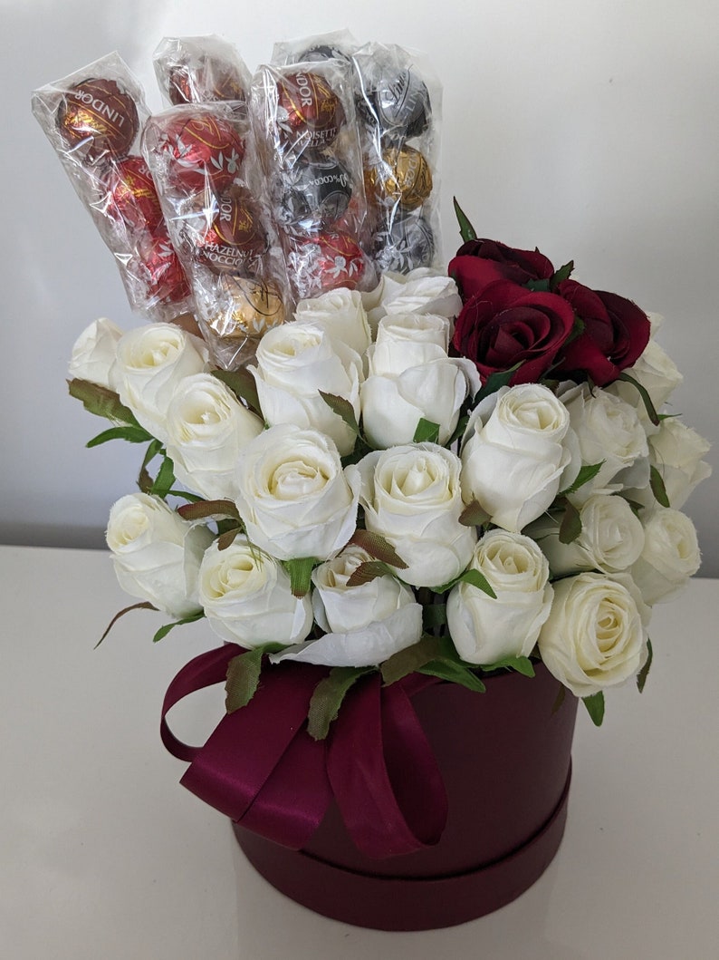 May include: A burgundy hat box filled with white roses and red roses. The box is tied with a burgundy ribbon. The box is filled with Lindor chocolates.