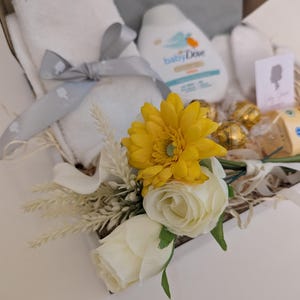 Puede incluir: Una cesta de regalo llena de art&iacute;culos para beb&eacute;s, que incluyen una toalla blanca, una botella de loci&oacute;n para beb&eacute;s, una flor amarilla y una flor blanca.