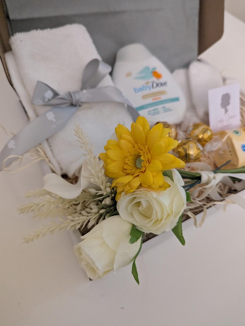 Puede incluir: Una cesta de regalo llena de art&iacute;culos para beb&eacute;s, que incluyen una toalla blanca, una botella de loci&oacute;n para beb&eacute;s, una flor amarilla y una flor blanca.