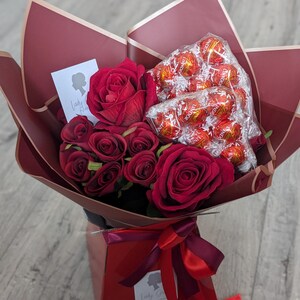 Puede incluir: Un ramo de rosas rojas y una caja de trufas de chocolate envueltas en papel rojo con una cinta roja. El ramo est&aacute; en una caja roja con una etiqueta blanca que dice "Lady G".