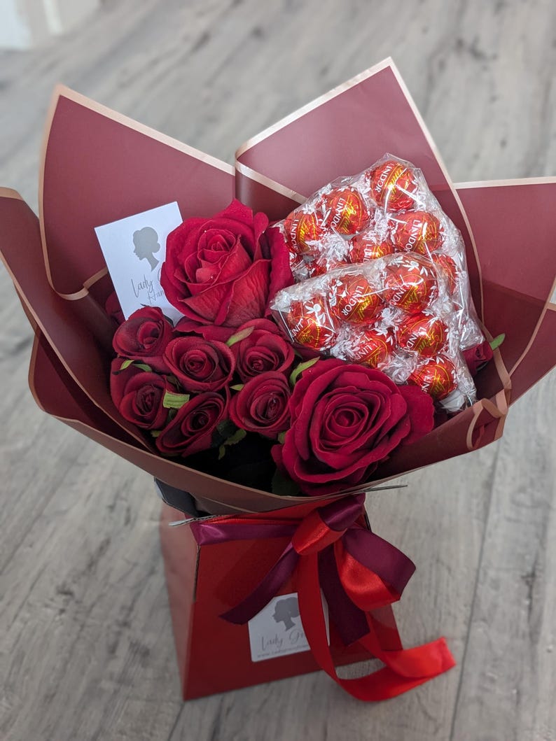Puede incluir: Un ramo de rosas rojas y una caja de trufas de chocolate envueltas en papel rojo con una cinta roja. El ramo est&aacute; en una caja roja con una etiqueta blanca que dice "Lady G".