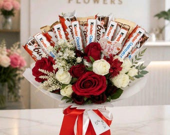 May include: A bouquet featuring red and white roses mixed with Kinder Bueno chocolate bars. The arrangement is wrapped in white paper and placed in a red gift bag with a red ribbon. A small white tag with text is attached to the red ribbon.
