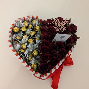 May include: A heart-shaped box filled with dark chocolate truffles and red roses. The box is decorated with white and red stripes and a red ribbon. The roses are arranged in a half-circle around the chocolates. A wooden sign with the text "Bride to Be" is placed on top of the roses.