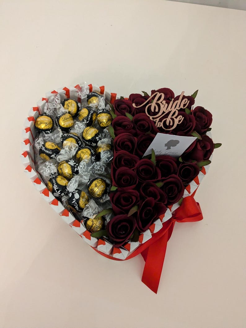 May include: A heart-shaped box filled with dark chocolate truffles and red roses. The box is decorated with white and red stripes and a red ribbon. The roses are arranged in a half-circle around the chocolates. A wooden sign with the text "Bride to Be" is placed on top of the roses.