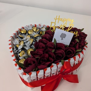 May include: A heart-shaped arrangement of chocolate sweets and red roses, surrounded by white chocolate bars. The arrangement is decorated with a red ribbon and a gold "Happy Birthday" topper.