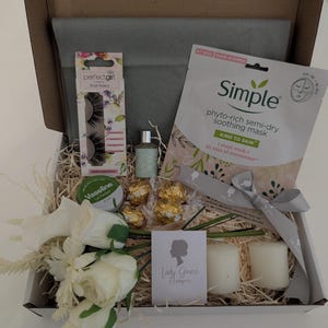 May include: A gift box filled with beauty and self-care items. Includes a Simple face mask, false eyelashes, Vaseline, a small perfume bottle, chocolates, candles, and a floral arrangement. The box is lined with tissue paper and decorative straw.
