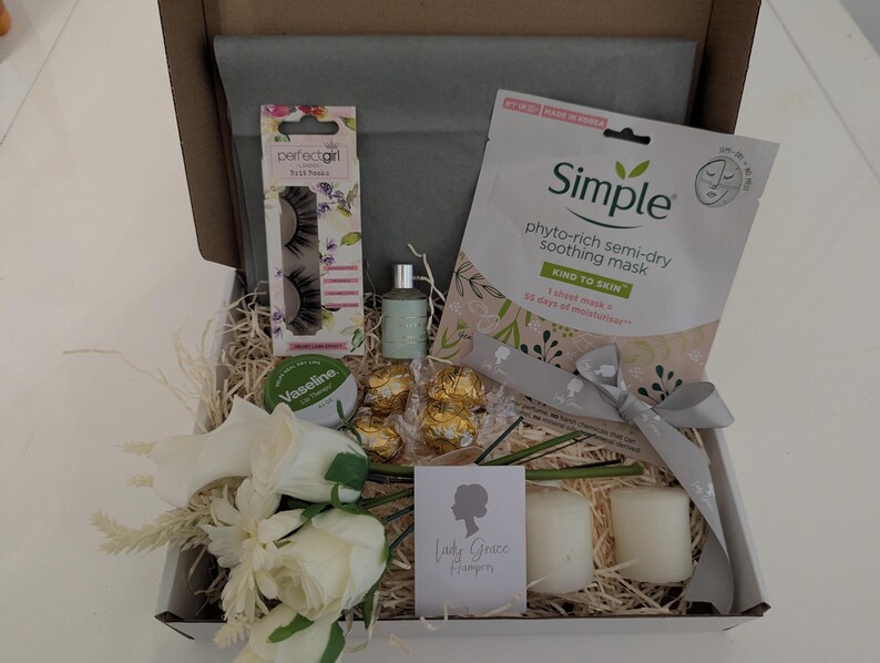 May include: A gift box filled with beauty and self-care items. Includes a Simple face mask, false eyelashes, Vaseline, a small perfume bottle, chocolates, candles, and a floral arrangement. The box is lined with tissue paper and decorative straw.