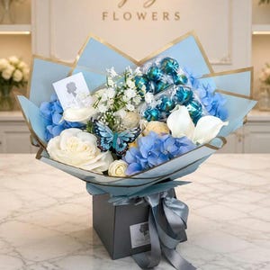 Pode incluir: Um buquê de rosas brancas, hortênsias azuis e gipsófila branca disposto em papel azul claro com bordas douradas. O arranjo inclui um ornamento de borboleta azul, esferas de chocolate azul-petróleo embaladas e um cartão branco com um logotipo.