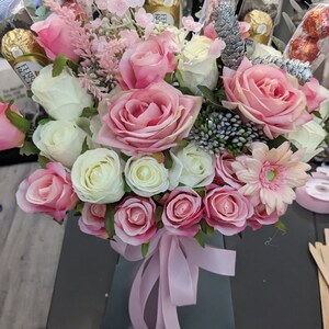 May include: A bouquet of pink and white roses, with a few pink carnations, in a gray box with a pink ribbon. The bouquet is decorated with chocolate wrapped in gold foil.