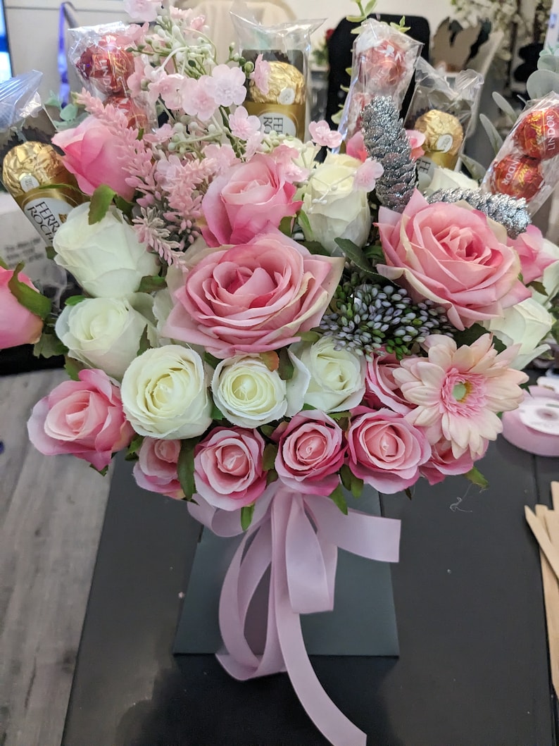 May include: A bouquet of pink and white roses, with a few pink carnations, in a gray box with a pink ribbon. The bouquet is decorated with chocolate wrapped in gold foil.