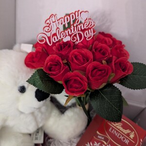 May include: A white teddy bear, a bouquet of red roses, and a box of Lindor milk chocolate truffles. The roses have a sign that says "Happy Valentine's Day".