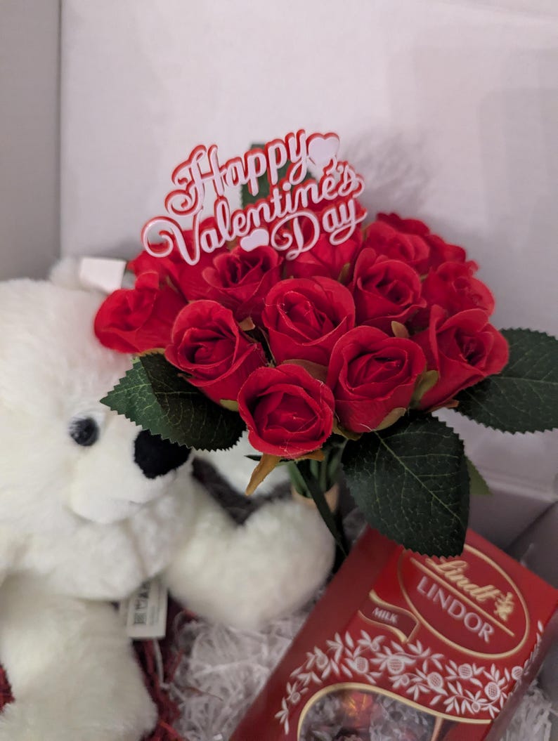 May include: A white teddy bear, a bouquet of red roses, and a box of Lindor milk chocolate truffles. The roses have a sign that says "Happy Valentine's Day".